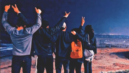 Devi Machete - During a chilly night, five CVYM volunteers embrace each other in a line as they face towards the border wall and the beach with their backs to the camera. November 19, 2019. Playas de Tijuana, México.